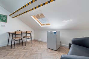 A small, modern room with a herringbone wood floor, a black sofa, a white refrigerator, a wooden table with chairs, and a skylight. - Top Floor Studio Apartment with Skylight & Fast WiFi in Ilkeston (Derbyshire)