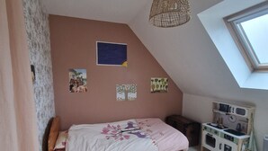 Ein Schlafzimmer mit schrägverdeckter Decke, einem Bett mit dekorativer Kopfteil, einem kleinen Holzschemel, einem an der Wand befestigten Regal mit einer Uhr und einem Oberlicht.