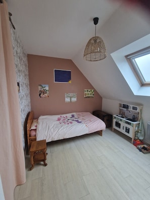 Ein Schlafzimmer mit schrägverdeckter Decke, einem Bett mit dekorativer Kopfteil, einem kleinen Holzschemel, einem an der Wand befestigten Regal mit einer Uhr und einem Oberlicht.