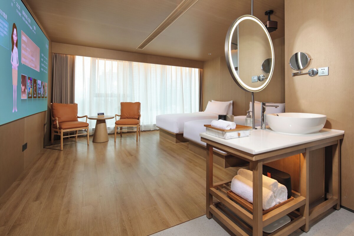 A hotel room with a large bed, a sitting area with two chairs, a round mirror, and a large white sink.