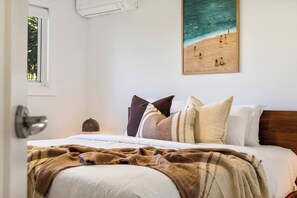 Ein Schlafzimmer mit einem Bett, Kissen, einer Decke, einem Bild von einer Strandansicht und einem Fenster mit Blick auf Bäume.