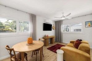 A modern living room with a round wooden table, a sofa, a TV mounted on the wall, and large windows with blinds. - Coolum Beach Coastal Retreat- Sleeps 10 (Coolum Beach)