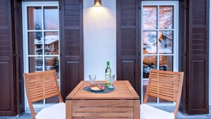 A wooden table with two chairs, a bottle of wine, and a plate of food on a balcony with wooden shutters and a wall-mounted light. - Grünstein Vacation Rental (Schönau am Königssee)