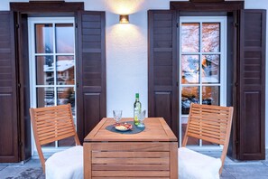 Ein Holztisch mit zwei Stühlen, einer Weinflasche und einem Teller Essen auf einem Balkon mit Holzläden und einer Wandleuchte.