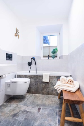 A compact bathroom with a white toilet, a bathtub, and a wooden bench with towels. - Grünstein Vacation Rental (Schönau am Königssee)