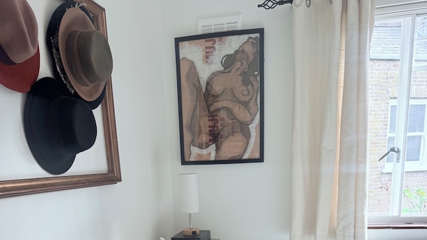 Een opgemaakt bed met een geweven witte hoes, een nachtkastje met een lamp, en een wand met lijstjes met hoeden en een foto.