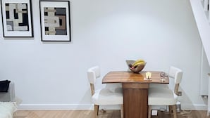 Een eethoek met een houten tafel, witte stoelen, een schaal fruit en een kaars. Twee ingelijste abstracte kunstwerken hangen aan de muur.
