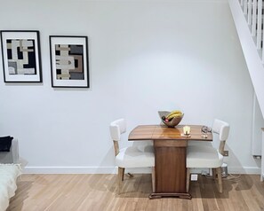 Ein Essbereich mit einem Holztisch, weißen Stühlen, einer Obstschüssel und einer Kerze. An der Wand befinden sich zwei gerahmte abstrakte Kunstwerke.