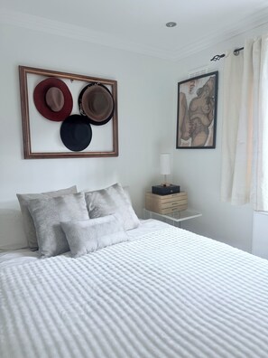 Quarto com cama, almofadas, mesa de cabeceira, luminária e quadros nas paredes.