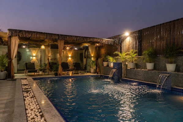 An indoor swimming pool with a waterfall feature, lounge chairs, and potted plants.