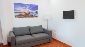 A grey sofa in a room with a painting of a sunset over rocks on the wall and a flat-screen TV mounted on the wall. - Terre di Monacella (Santa Venerina)