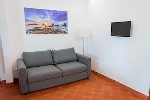 A grey sofa in a room with a painting of a sunset over rocks on the wall and a flat-screen TV mounted on the wall. - Terre di Monacella (Santa Venerina)