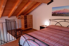 A bedroom with a sloped ceiling, a bed with a striped bedspread, a wooden cabinet, and a picture of a mountain on the wall. - Terre di Monacella (Santa Venerina)