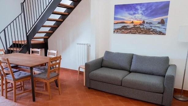 A living room with a sofa, a dining table with chairs, a staircase, and a painting of a sunset. - Terre di Monacella (Santa Venerina)