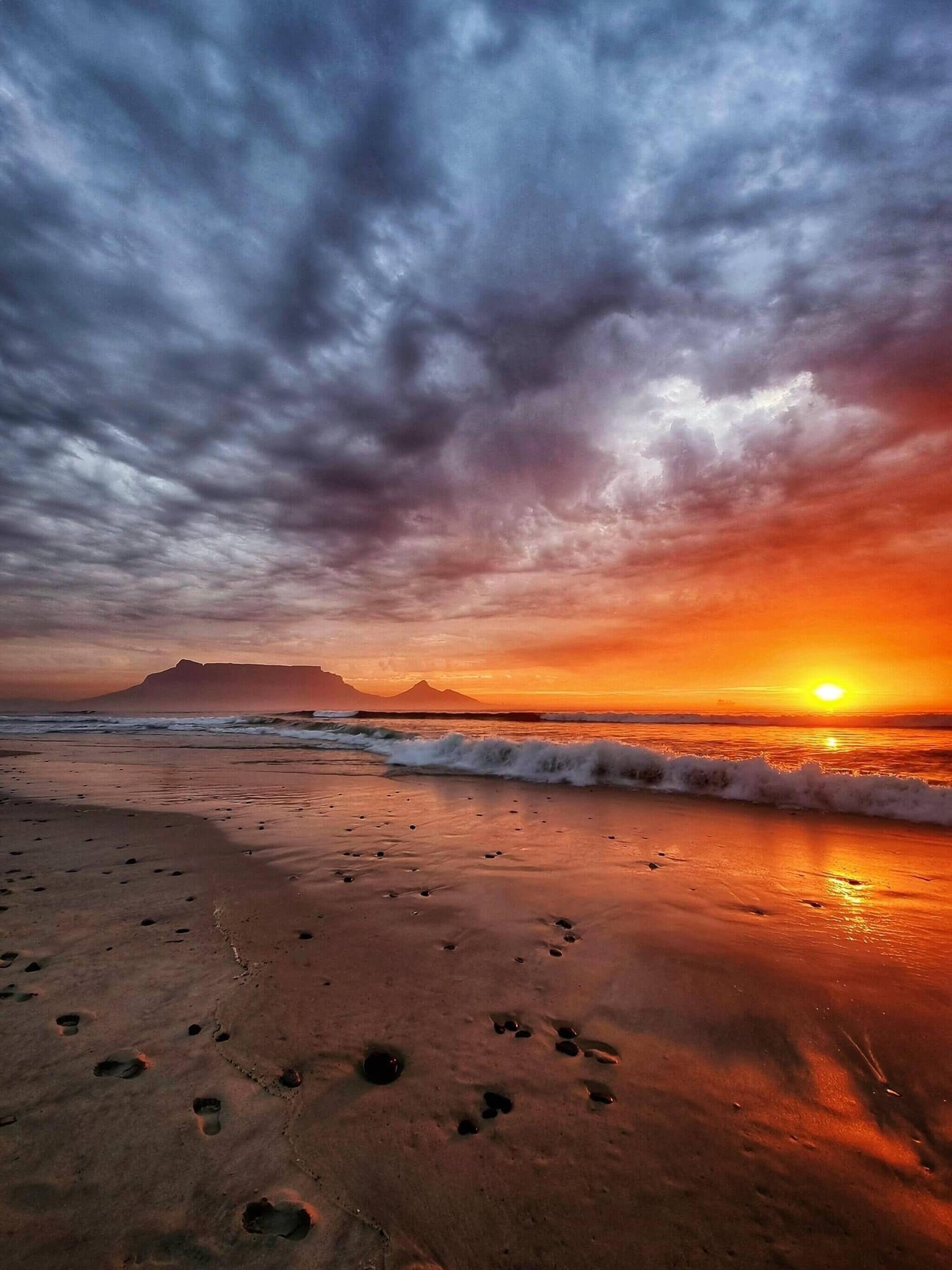 A beach with footprints leading towards the ocean, waves approaching the shore, and a sunset in the background.