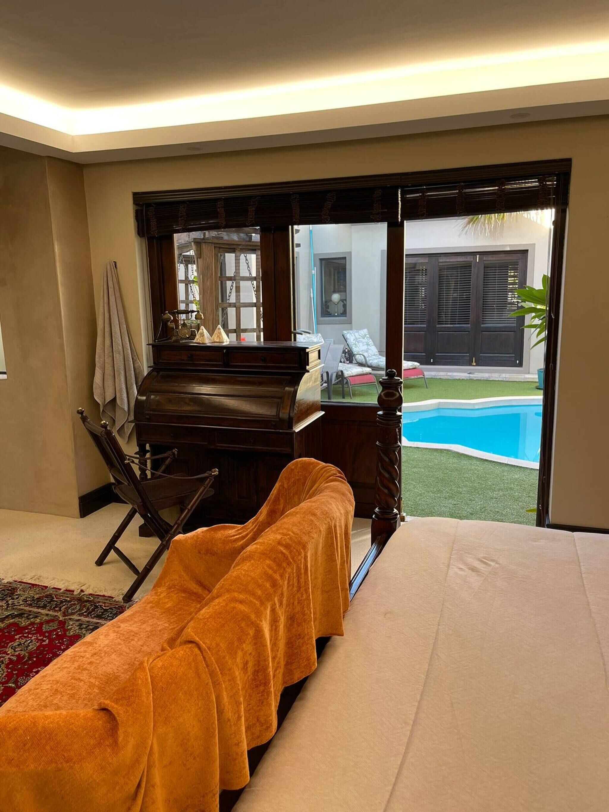 A bedroom with a bed, a wooden desk, a chair, a massage table, and a view of a swimming pool.