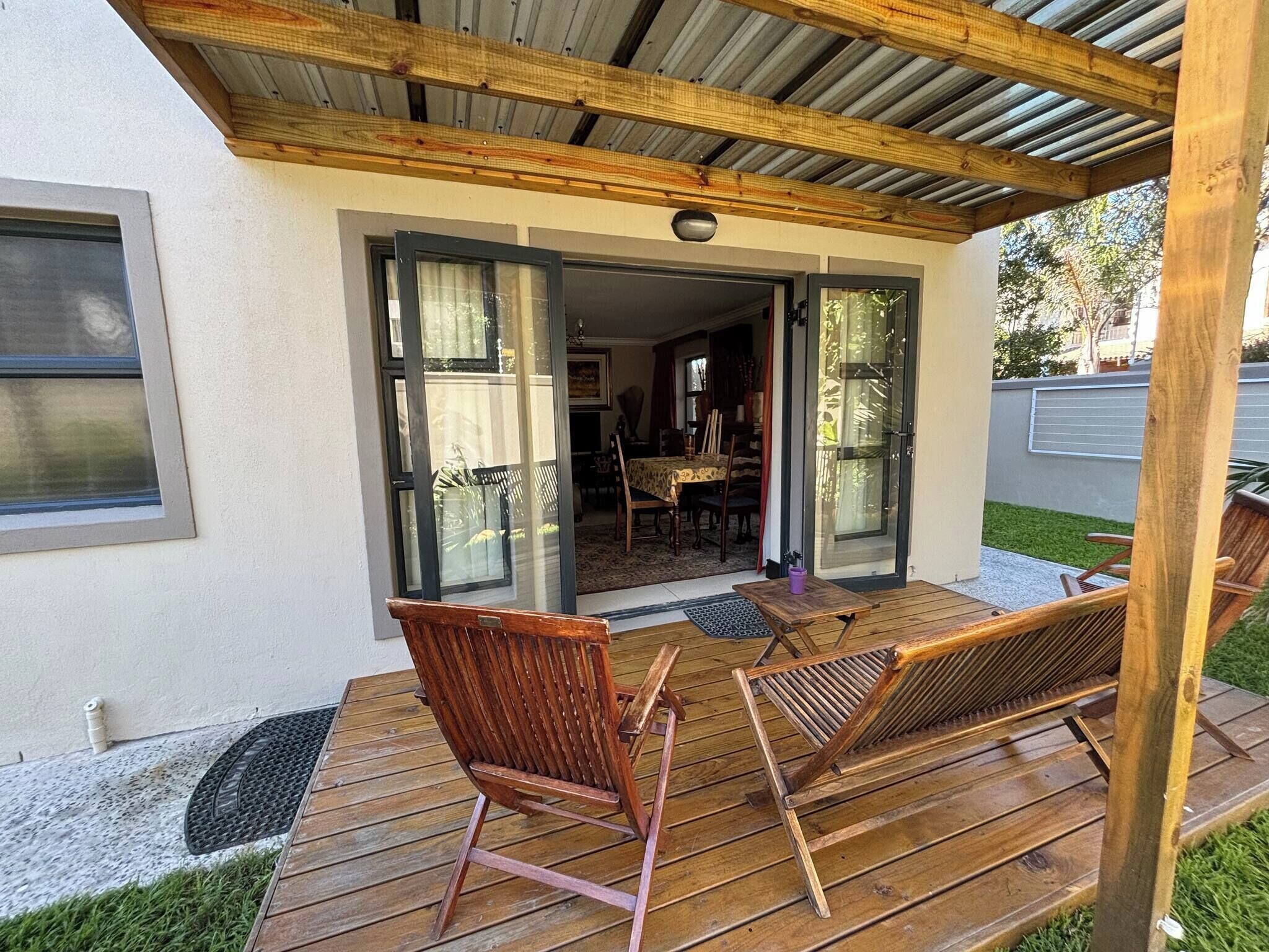 An outdoor wooden deck with two wooden chairs and a table, a covered patio area with a metal roof, and a glass door leading to an interior dining area.