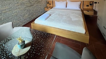 Un dormitorio con cama, una mesita de noche, una silla, una pequeña mesa con una planta, un aire acondicionado de pared y un cuadro en la pared.