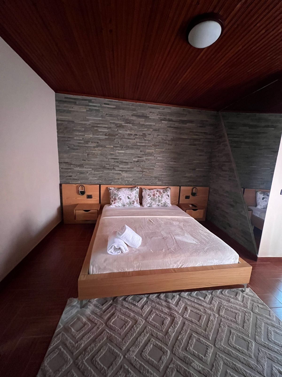 A bedroom with a wooden bed, a stone-patterned wall, and a ceiling light fixture.
