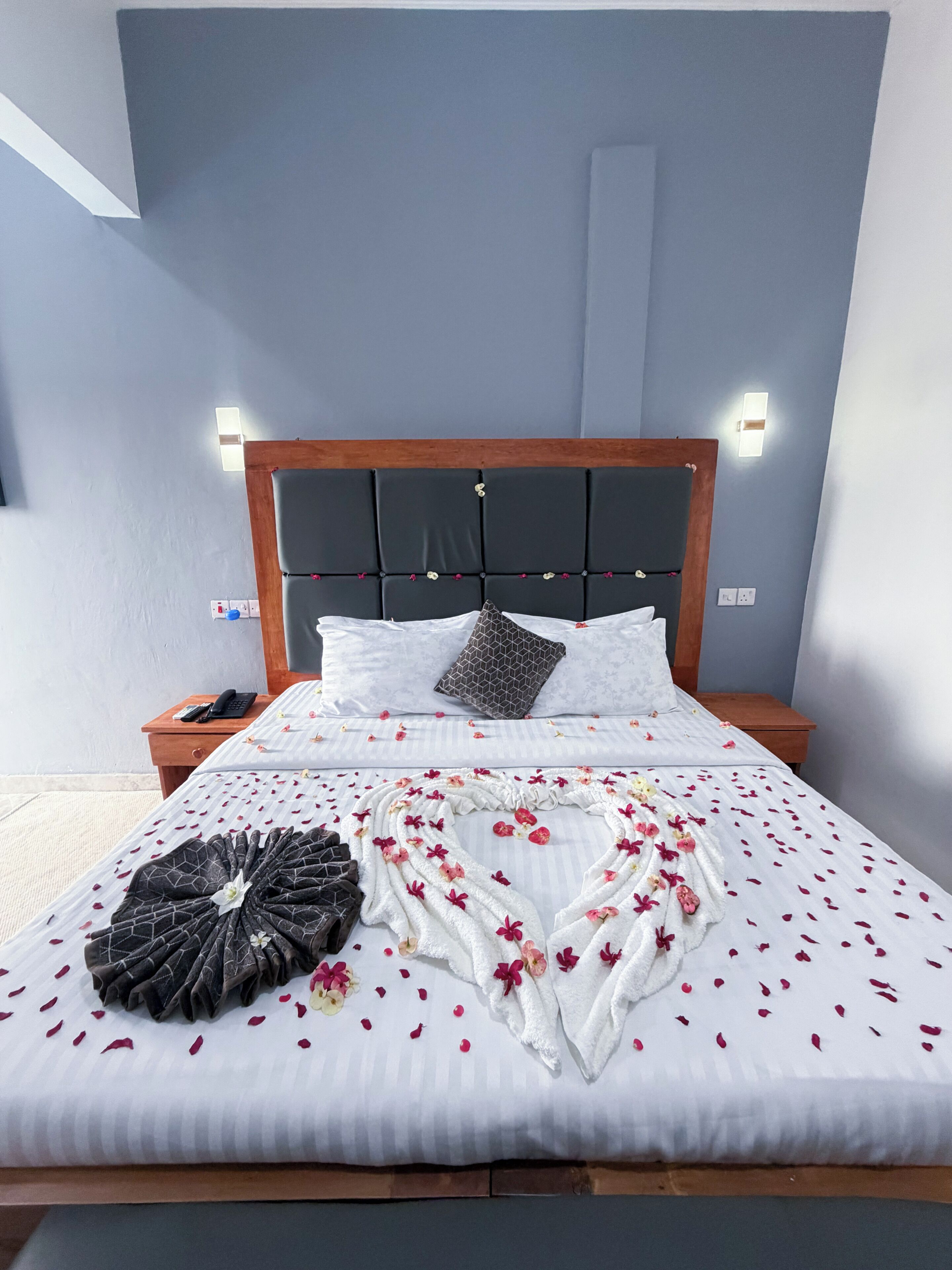 A neatly made bed with a heart-shaped arrangement of flowers and a blanket, a wooden headboard, and bedside lamps.