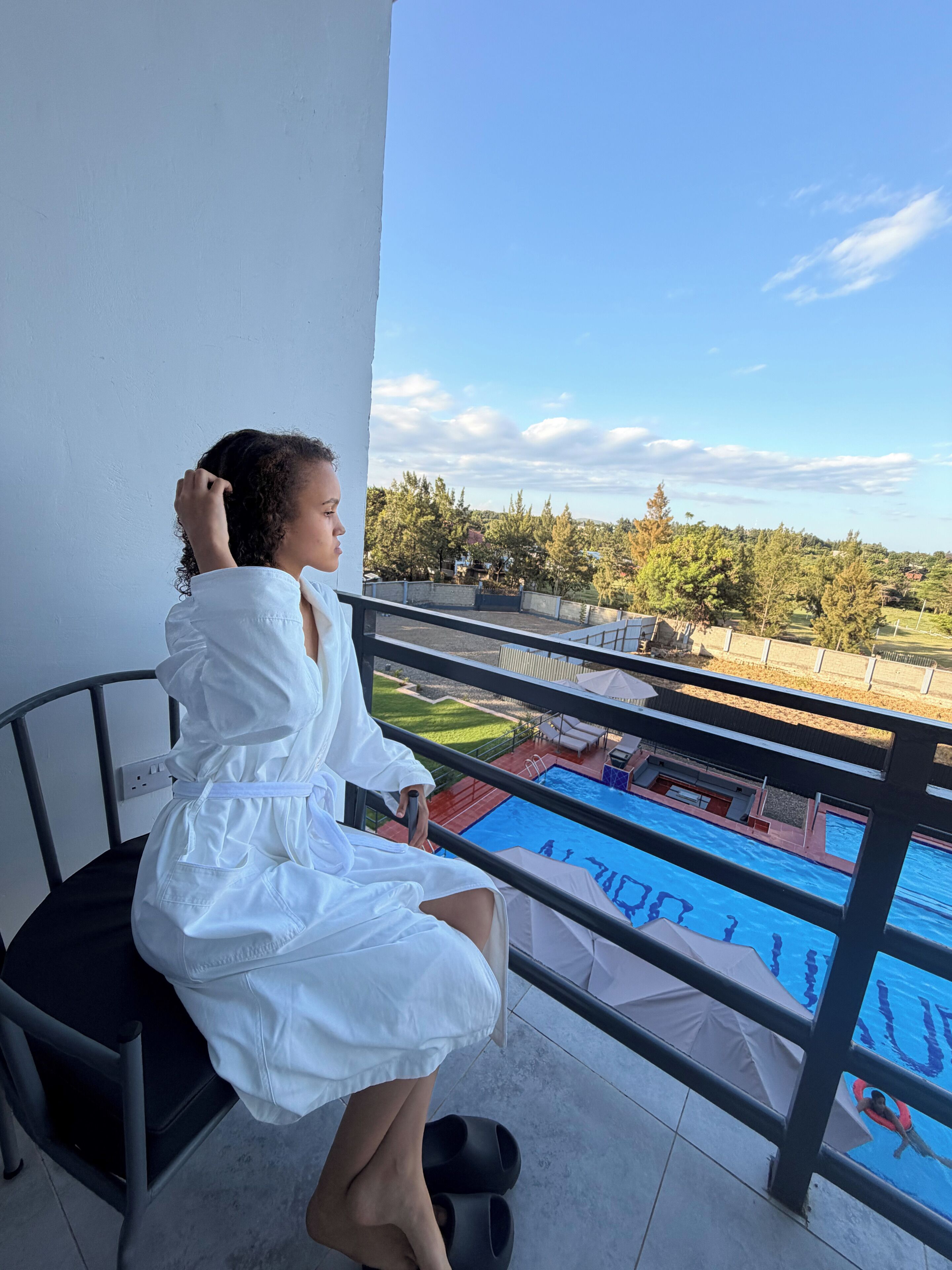 Una persona sentada en una silla de balcón con vista a una piscina, árboles y cielo despejado.