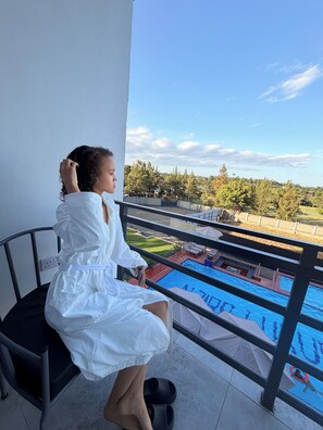 Una persona sentada en una silla de balcón con vista a una piscina, árboles y cielo despejado.