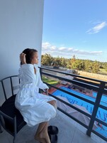 Una persona sentada en una silla de balcón con vista a una piscina, árboles y cielo despejado.