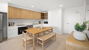 A modern kitchen with white cabinetry, a wooden dining table, and a bench. - Poolside 2-bed Apartment at Lake Kawana (Sunshine Coast)