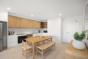A modern kitchen with white cabinetry, a wooden dining table, and a bench. - Poolside 2-bed Apartment at Lake Kawana (Sunshine Coast)
