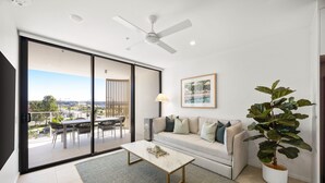 A modern living room with a sliding glass door leading to an outdoor area, a ceiling fan, a sofa with cushions, a coffee table, and a potted plant. - Poolside 2-bed Apartment at Lake Kawana (Sunshine Coast)