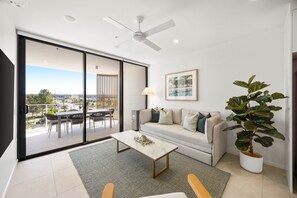 A modern living room with a sliding glass door leading to an outdoor area, a ceiling fan, a sofa with cushions, a coffee table, and a potted plant. - Poolside 2-bed Apartment at Lake Kawana (Sunshine Coast)