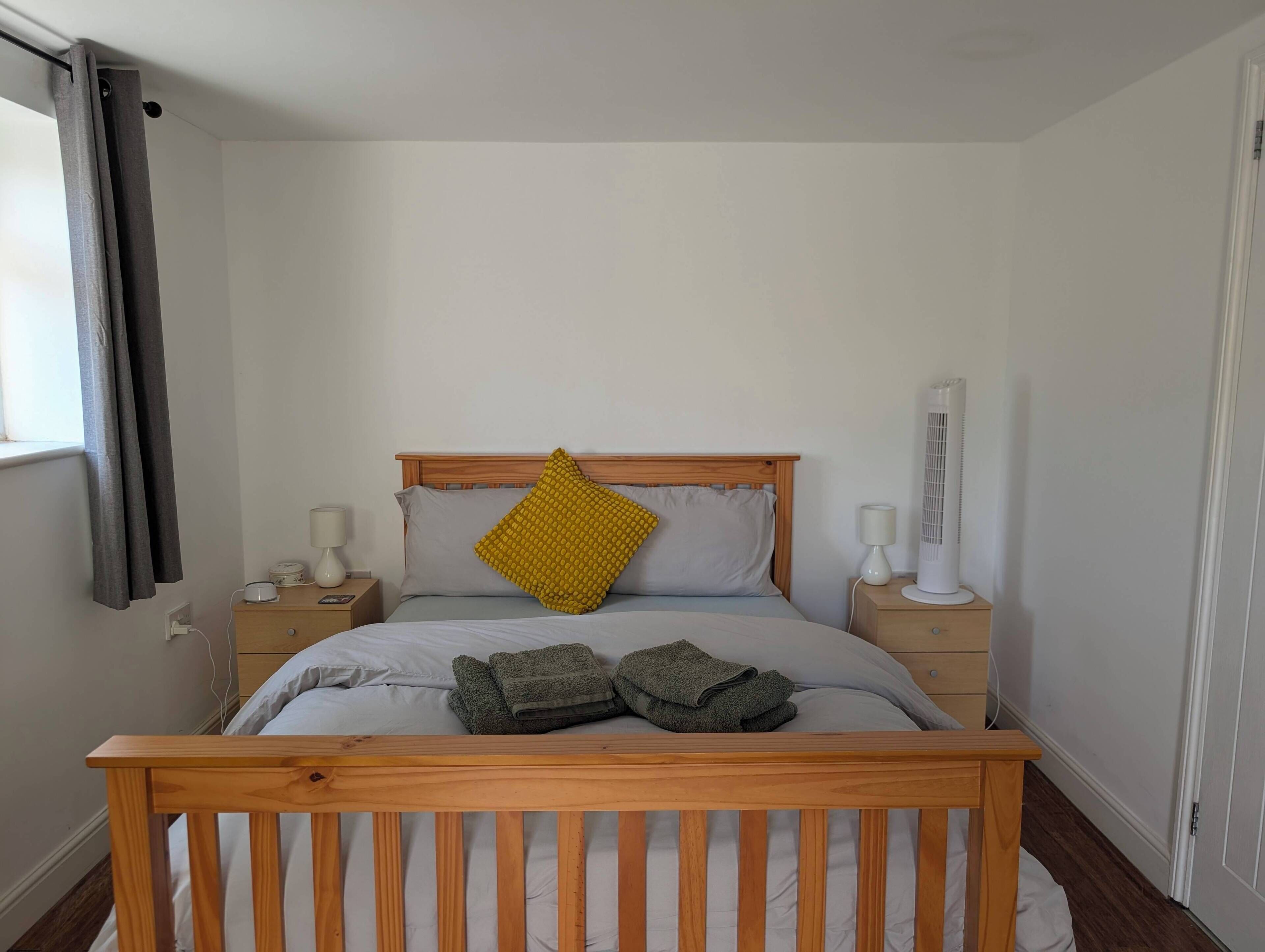 A bedroom with a wooden bed frame, a single bed with grey bedding, a yellow pillow, two bedside tables with lamps, and a white radiator.