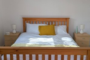 Una cama con cabecera de madera, ropa de cama blanca y un almohadón amarillo. Hay dos mesitas de noche con lámparas y un adorno decorativo en cada una.