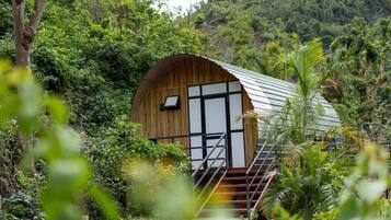 A wooden cabin with a metal roof, surrounded by dense greenery and a staircase leading up to the entrance.
