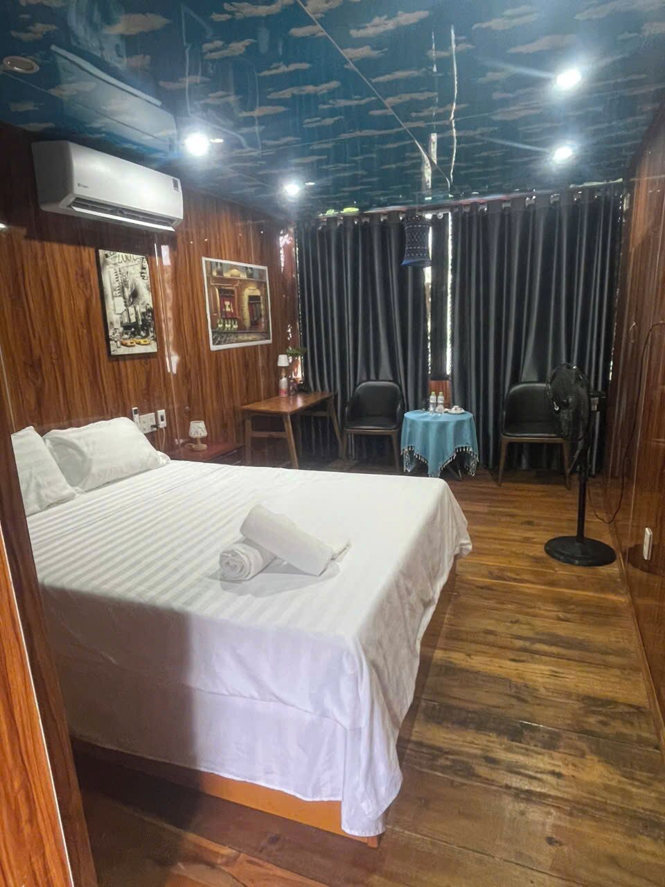 A hotel room with a large bed, wooden flooring, a ceiling fan, a table with chairs, and a window with curtains.