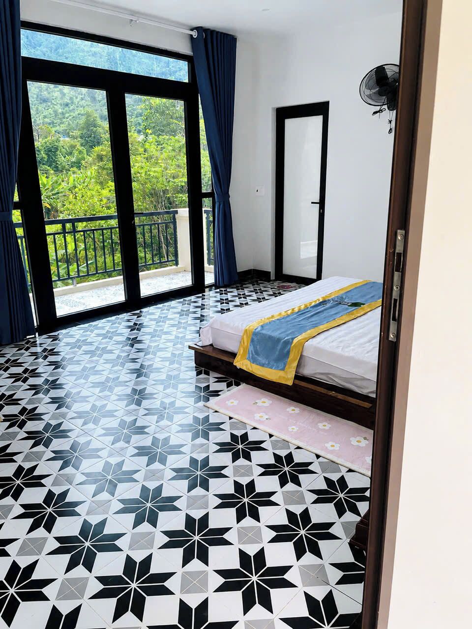 A room with a bed, a fan, and a view of greenery through large windows.