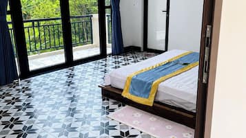 A room with a bed, a fan, and a view of greenery through large windows.