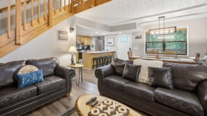 A modern living room with leather sofas, a wooden staircase, a kitchen in the background, and a large window with blinds. - Bear Run Getaway | Near Downtown Gatlinburg (Gatlinburg)