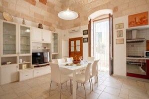 A dining area with a white table and chairs, a television, and a kitchen with a stove and microwave.