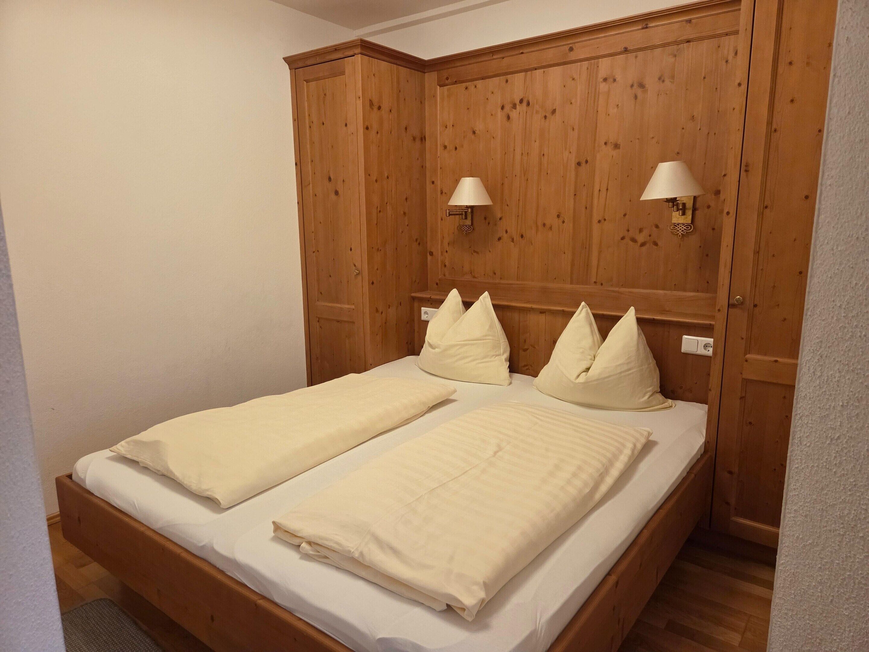 Een tweepersoonsbed met twee kussens, een houten hoofdbord met wandlampen en een houten garderobe.