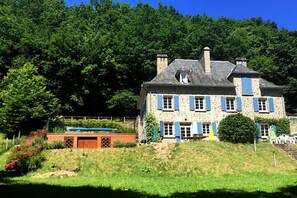 Un bâtiment en pierre doté de nombreuses fenêtres et d’une cheminée, entouré d’une végétation luxuriante.