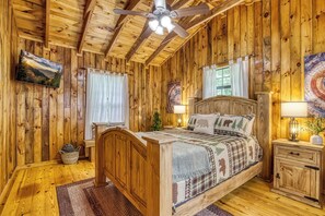 Ein Schlafzimmer einer Holzhütte mit einem Bett, Nachttischen, einem Deckenventilator und einem Fenster mit Vorhängen.