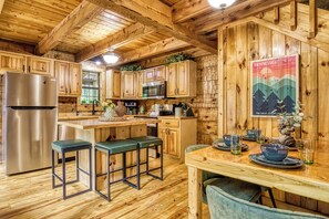 Eine rustikale Kücheneinrichtung im Chalet-Stil mit Holzausstattung, einem Edelstahlkühlschrank, einer Mikrowelle und einem Essbereich mit Hockern und Tisch.