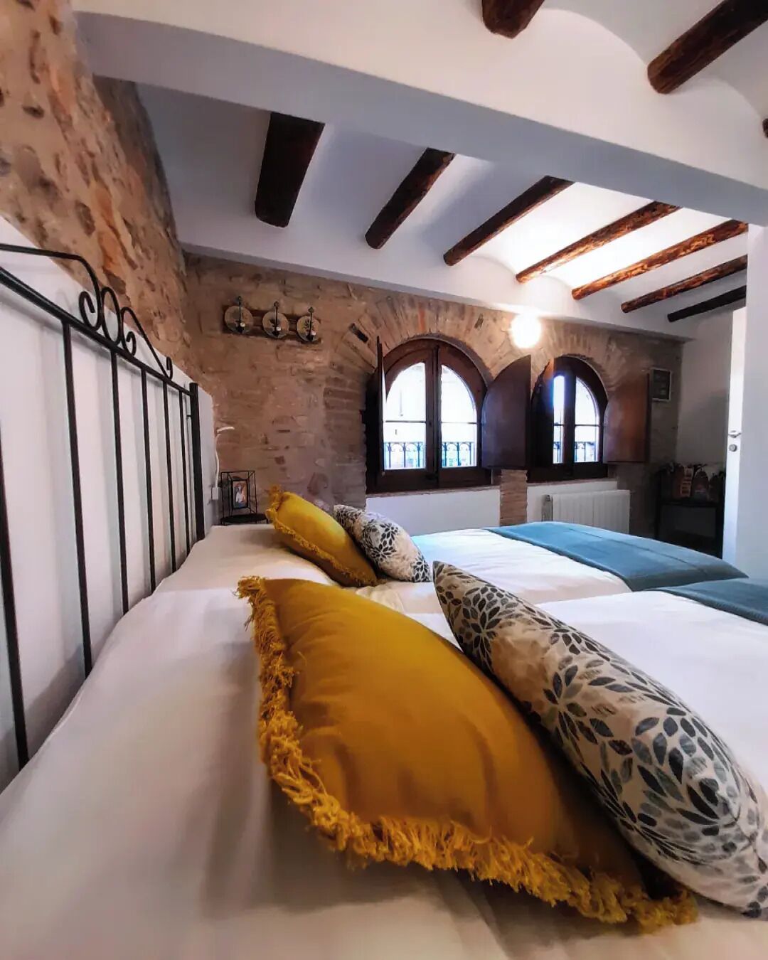 A bedroom with a stone wall, a bed with white bedding and patterned pillows, and a view of a balcony through an arched window.