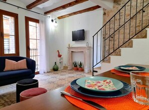 A living room with a sofa, ottoman, and a dining table set with plates and a knife. A fireplace and a television are visible in the background.