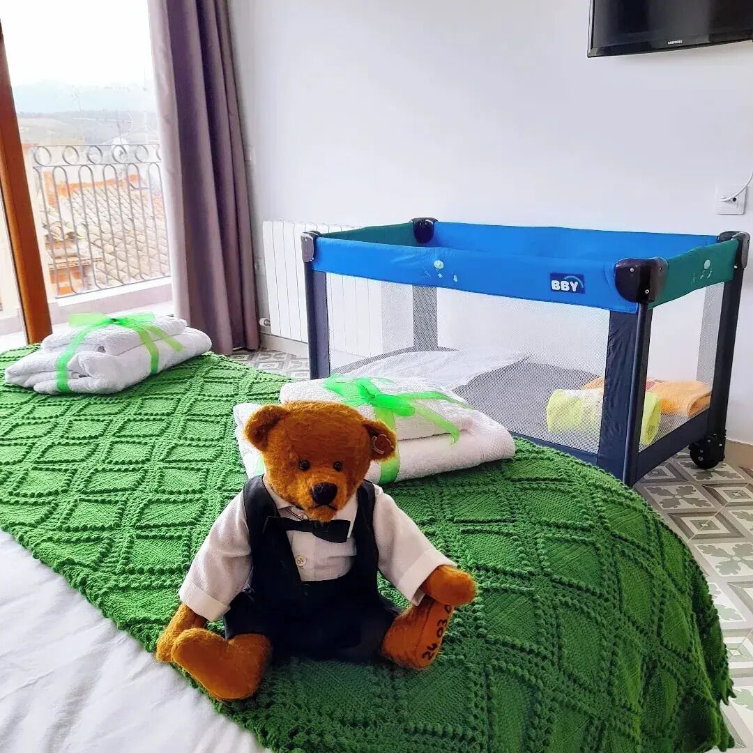 A room with a bed, a green knitted blanket, a baby playpen, and a teddy bear.