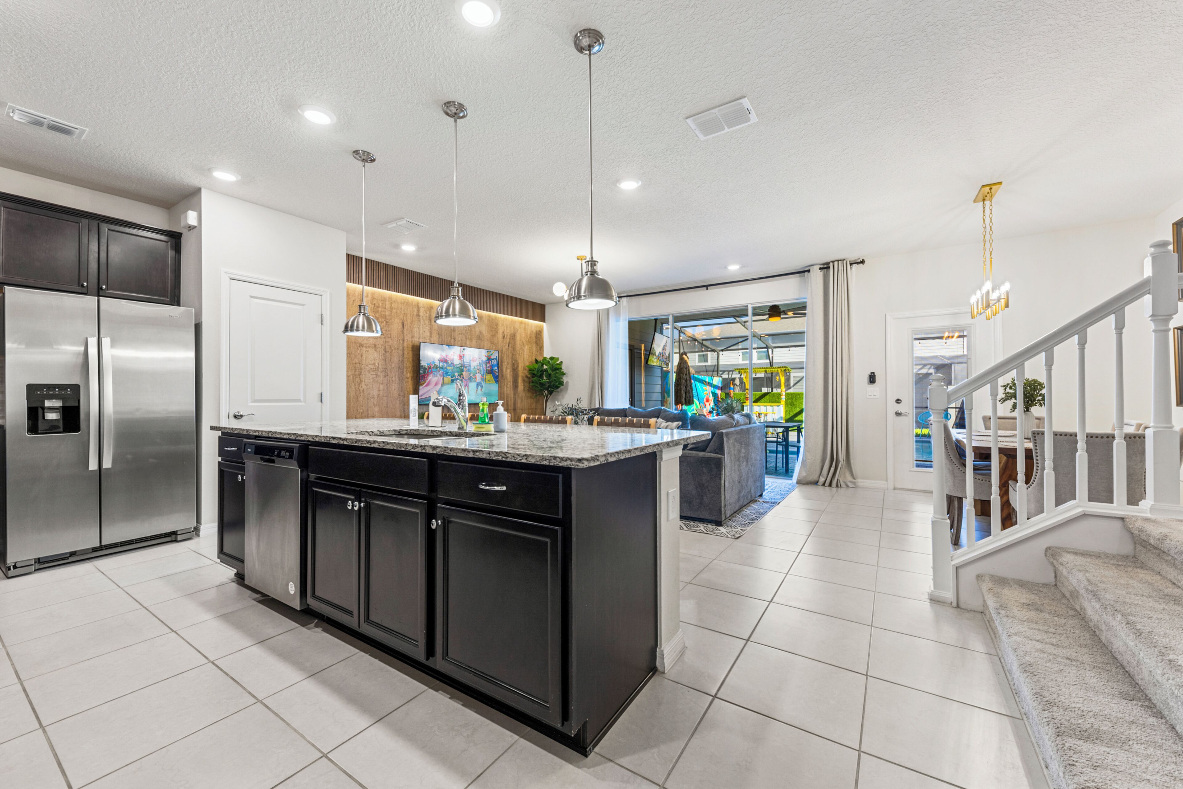A modern kitchen with a central island, stainless steel appliances, pendant lighting, and a view into a living area with a staircase.
