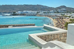 A coastal resort with a clear pool, beach, and white buildings. - Beachfront Mykon Villa – Your Relaxing Getaway in Platys Gialos (mykonos)