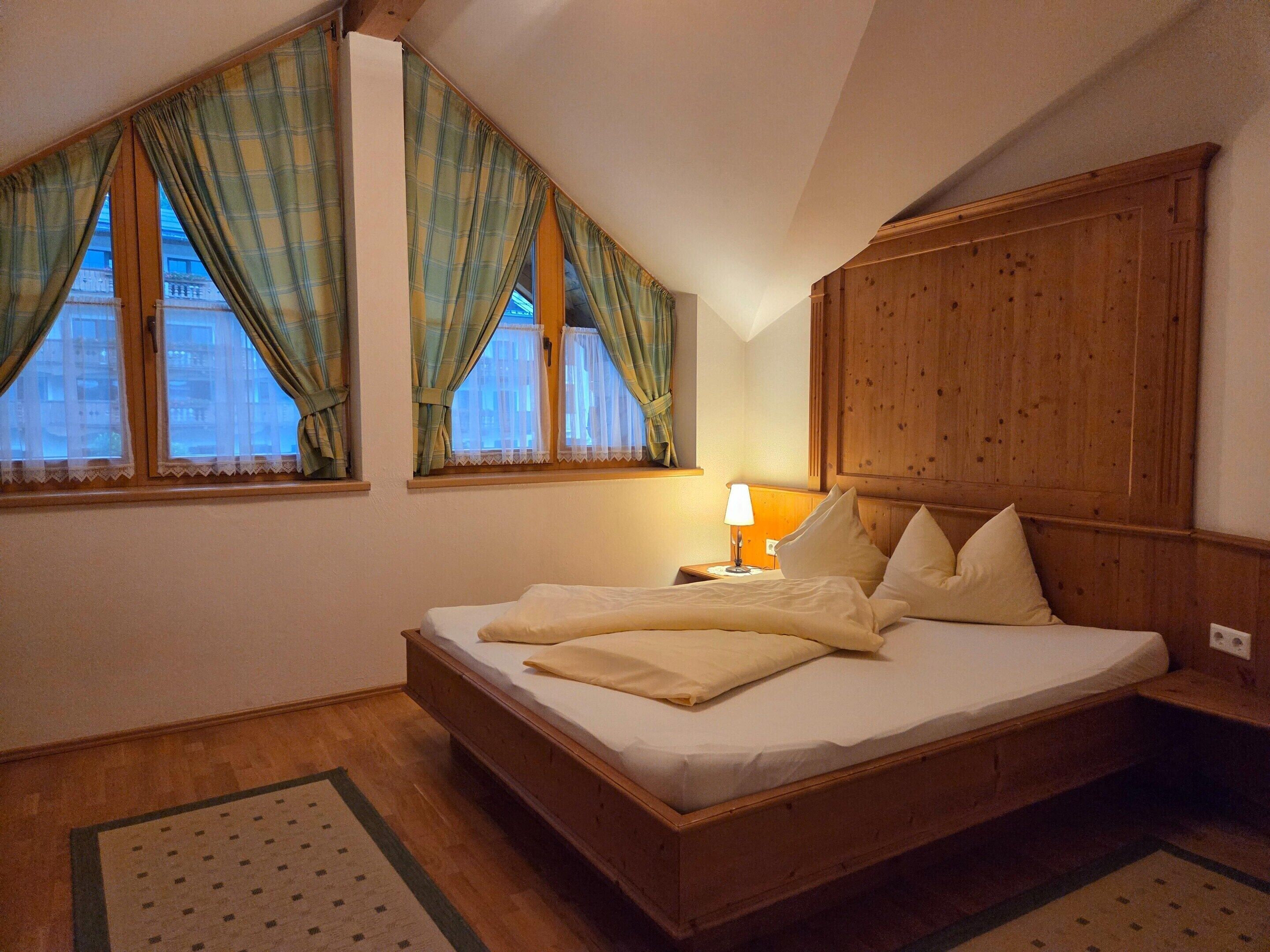 A bedroom with a wooden bed, two windows with curtains, a nightstand with a lamp, and a wooden headboard.