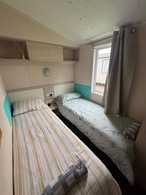 Una habitación pequeña y funcional con dos camas individuales, una ventana con cortinas y un estante en la pared.